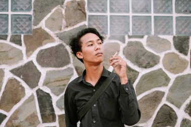 Genç adam dışarıda sigara içiyor, arka planda taş var, derin derin bakıyor.