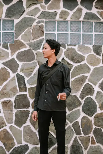 Elinde sigara tutarken taş bir duvarın yanında duran siyah elbiseli genç adam.