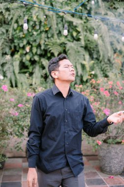 Yeşil bitkilerle çevrili bir adam dışarıda sigara içiyor. Düşünceli ve düşünceli görünüyor..