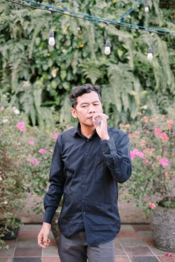 Dalgın bir adam yemyeşil bir bahçe ortamında bitkisel sigara içiyor. Rahatlama ve huzur dolu anlar.