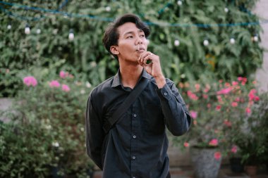 Elinde sigara tutan genç bir adam, açık havada yemyeşil bir alanda derin derin düşünüyor. Düşünceli bir ifadeyle sıradan bir tarz.