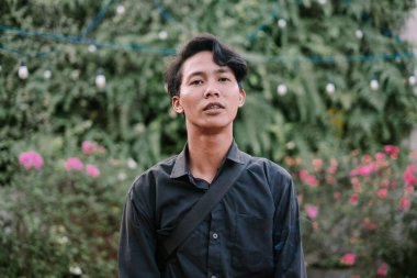 Asyalı genç bir adamın otantik portresi. Pembe flora ile yeşil bir bahçe arkaplanının ortasında güvenle duruyor.