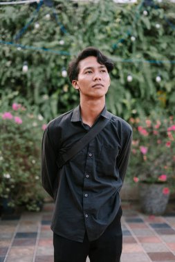 Düşünceli bir Asyalı genç adam, yemyeşil ve çiçeklerle çevrili, günlük şık bir kıyafetle dışarıda poz veriyor..