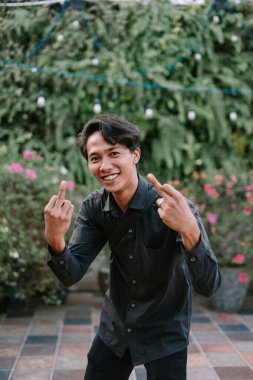 Gülümseyen genç bir adam dışarıda, yemyeşil bir bahçe ortamında poz verirken şakayla karışık saldırgan bir jest yapıyor. Modern tutumun gelişigüzel ve meydan okuyan bir görüntüsü..