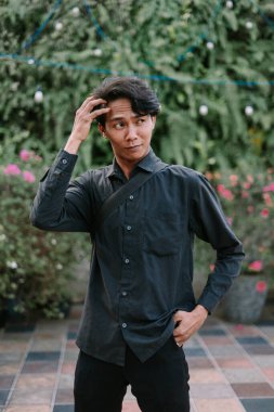Dışarıda duran dalgın görünümlü Asyalı bir adam. Siyah bir gömlek giyiyor, yemyeşil bir bahçede öylesine poz veriyor.
