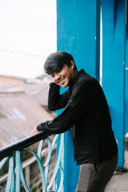 Parmaklıklara yaslanmış neşeli Asyalı bir adam açık hava ortamında samimi bir gülümsemeyle. Pozitif duygu ve gençlik yaşam tarzı, kentsel çevre