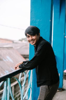 Mavi bir balkonda dostça gülümseyen genç bir adam. Siyah bir gömlek giyiyor ve rahatlıyor..