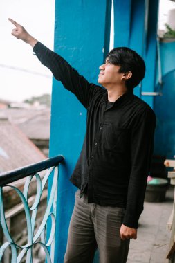 Parmakla gökyüzünü gösteren bir adam, canlı mavi bir arka planı olan bir evin verandasında duruyor.