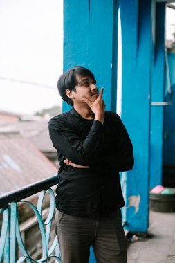 Gündelik giysiler içinde, balkonda duran genç bir adam. Düşünceli bir şekilde yukarı bakıyor, belki de bir fikir düşünüyor.