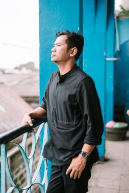 Asyalı bir adam mavi bir balkonda duruyor. Düşüncelere dalmış, geleceği düşünüyor..