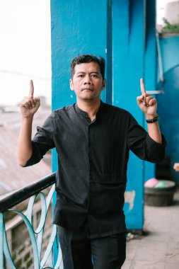 Balkonda dikilip kameraya ciddi bir ifadeyle bakan düşünceli bir adam.