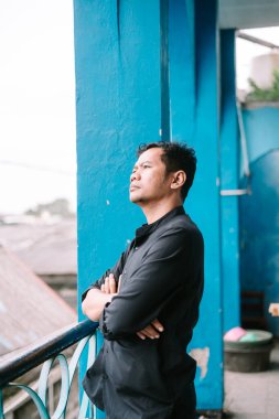 Asyalı bir adam parlak mavi bir balkonda düşünceli bir şekilde uzağa bakar. Kollarını kavuşturup dalgın dalgın bakıyor..