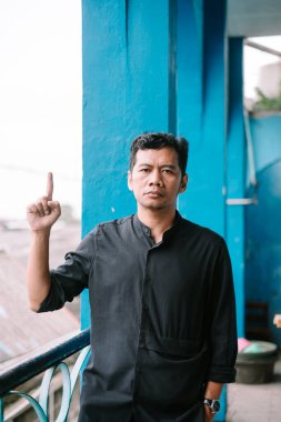 Düşünceli Asyalı bir adam işaret parmağını yukarı kaldırıp mavi desenli bir duvarın önündeki kameraya bakıyor..