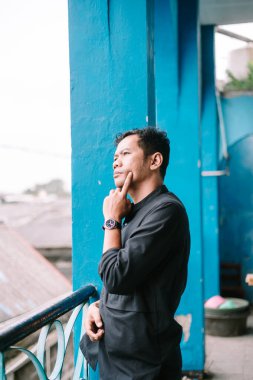 Derin düşüncelere dalmış düşünceli Asyalı bir adam başka tarafa bakıyor. Mavi duvara yaslanmış düşünceli bir ifade.