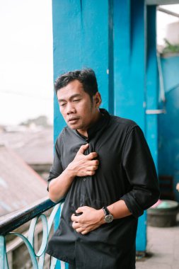 Endişeli bir adam balkonda göğsünü rahatsız ediyor. Ağrısı ya da sıkıntısı var gibi görünüyor, muhtemelen bir sağlık sorunu var.