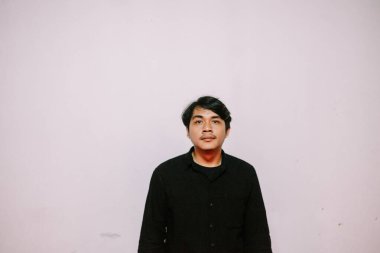 Yüzünde ciddi bir ifade olan, açık renkli bir arkaplana karşı siyah gömlek giyen genç bir adamın portresi. Tarafsız ve temiz estetik