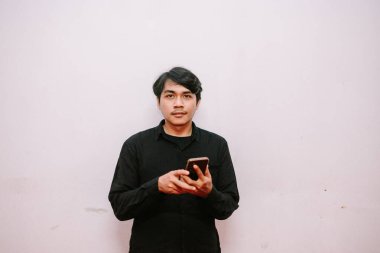 Akıllı telefonlu genç adam, günlük siyah gömlekli pembe arka planda kameraya bakıyor.