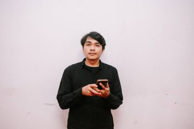 Siyah gömlekli genç bir adam akıllı telefonunu düz bir duvara karşı kullanıyor. O, tesadüfi bir şekilde doğrudan kameraya bakıyor..