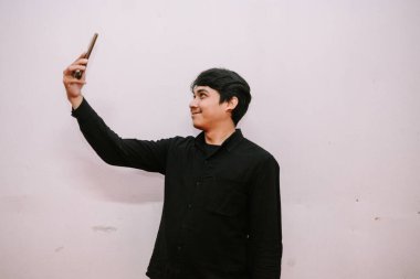 Siyah gömlekli genç adam, beyaz arka planda izole edilmiş akıllı telefonuyla selfie çekiyor.