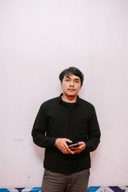 Beyaz arka plan önünde cep telefonu tutan genç bir adamın portresi. Kameraya tarafsız bir ifadeyle bakıyor..