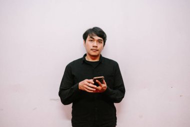 Koyu renk tişörtlü, telefon duvara dayalı bir adam. Elinde akıllı bir telefon var ve kameraya bakıyor..