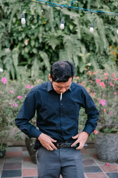 Dışarıda sigara içen bir adam, yeşil ve yumuşak dekoratif ışıklarla çevrili. Kemerini ayarlıyor..