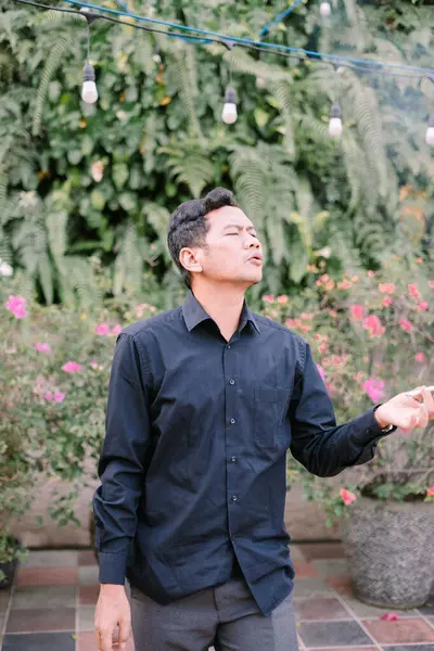 Yeşil bitkilerle çevrili bir adam dışarıda sigara içiyor. Düşünceli ve düşünceli görünüyor..