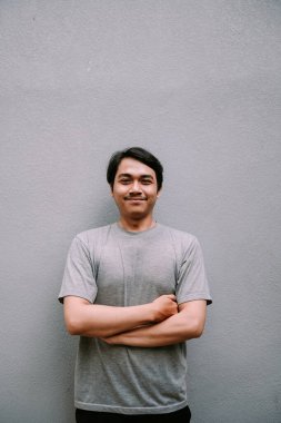 Gri bir arkaplanda kollarını kavuşturmuş gülümseyen Asyalı bir adam, kendine güveni ve sıradan bir yaşam tarzı yayıyor.