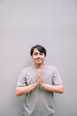 Tarafsız gri bir arkaplan önünde ellerini sallayarak selam veren genç bir adam. Hoş geldin ve saygı ifadesi.