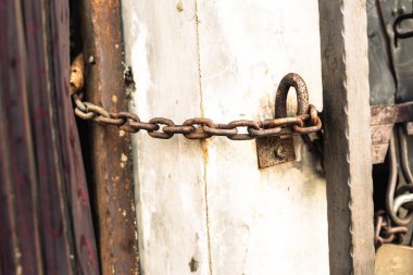 Metal bir kapıyı beton bir duvara bağlayan paslı metal bir zincirin yakın çekimi. Zincir çok yıpranmış ve yaş ve kullanım belirtileri gösteriyor..