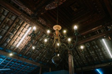 Karmaşık bir şekilde tasarlanmış koyu ahşap tavandan sarkıtılmış süslü bir avize. Tavanda ayrıntılı oymalar ve geleneksel mimari stil sergileniyor..