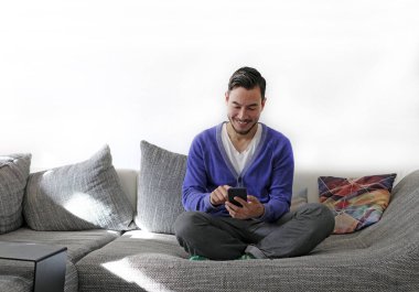 Kanepede otururken akıllı telefonu tutan gülümseyen adam.