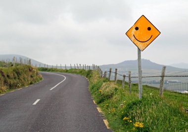 İyimserlik - Tepe manzarasında bir eğrinin önünde gülen yüzlü sarı yol işareti