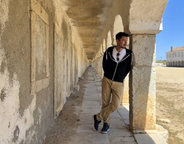 Havacı güneş gözlüklü bir adam gömleğinin yakasına sıkışmış terk edilmiş bir binanın sütununa yaslanmış.