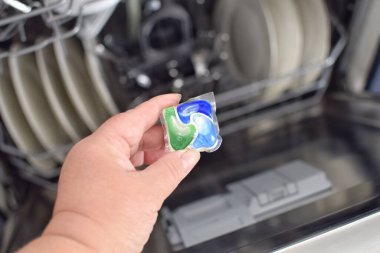 Bir kadın bulaşık makinesinin arka planında kirli bulaşıklarla deterjan kapsülü tutuyor.