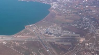 Flying above runway extended to sea, looking to the right of the frame. Passenger window view aboard airplane cruising at low altitude above SKG airport Thessaloniki, Greece.
