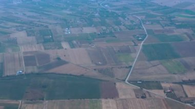 Flying above Mediterranean terrain with fields and scattered houses. Passenger window view aboard an airplane cruising, looking to the left of the frame in Greece.