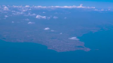 Flying above coastal terrain with clouds on the horizon. Passenger window view aboard an airplane cruising at high altitude, looking to the left of the frame.