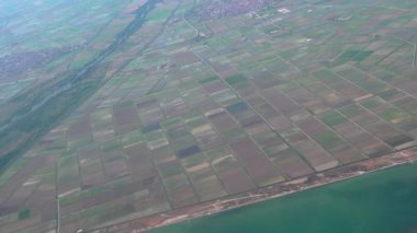 Flying above Mediterranean terrain with fields, a river and small settlements. Passenger window view aboard an airplane cruising, looking to the right of the frame in Greece.
