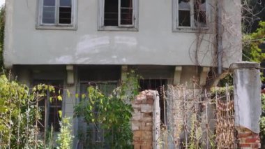 Day pan to an old low-rise stone-built damaged house with rusty iron railings and high-rise green vegetation around in Florina, Greece.