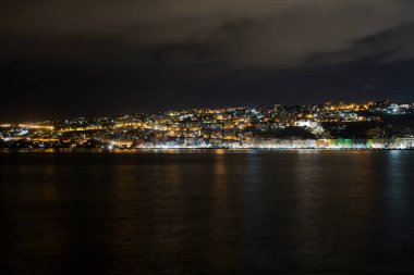 Gece boyunca bulutlu bir gökyüzünün altında binalar ve ışıklarla kaplı bir şehri çevreleyen deniz.