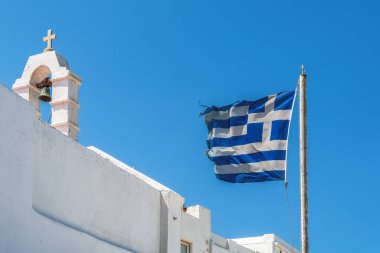 Beyaz bir Ortodoks kilisesi, çan kulesi ve Hıristiyan haçı ile birlikte Mykonos Adası 'nda güçlü bir rüzgârın estiği gökyüzü arka planında dalgalanan Yunan bayrağı.