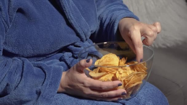 一个涂有光滑指甲的女人坐在沙发上 吃着一碗清澈的薯片 穿着蓝色长袍 散发着舒适和放松的气息 看电视前的肥胖概念 — 图库视频影像