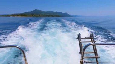 Yelkenli gemi gününün arka tarafında mavi denize bakan alçak dalgalar ve ufuk boyunca sürat teknesinin bıraktığı köpüklü su izleriyle kara ya da ada görülebilir..