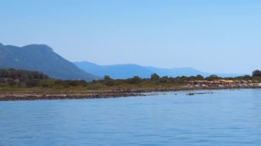 Yelkenli gemi manzarası yeşil bitkilerle kaplı kayalık bir sahil şeridine bakıyor ve uzak uçlarda tanımlanamayan banyoları olan bir sahil barı var..