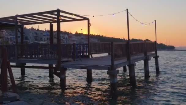 セファロニアギリシャのイオニア島のArgostorion町で日没時に海の上の木製デッキに椅子やテーブルを持つ空のウォーターフロントの居酒屋 — ストック動画