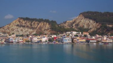 Zakynthos, Yunanistan - 11 Temmuz 2021: İyon adası deniz manzarası alçak rıhtımlı binalar. Sakin denizin yanındaki kayalık tepenin altındaki ev ve mağazaların manzarası..