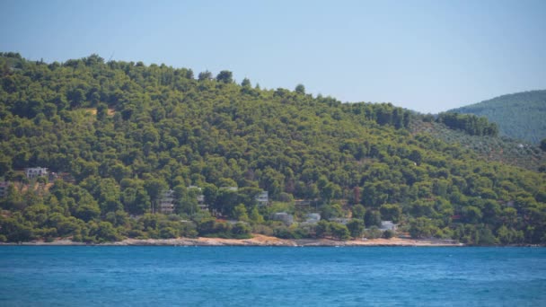 低層の散らばったビーチハウスは 一部はギリシャの穏やかな青い海で丘の上の緑の木と森の周りに隠れています — ストック動画