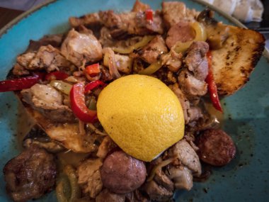Tavuk, domuz eti ve sosis de dahil olmak üzere pişirilmiş et parçaları kırmızı ve yeşil biberli, mantarlı ve soğanlı ve dilimlenmiş limonlu bir tabakta servis ediliyor..