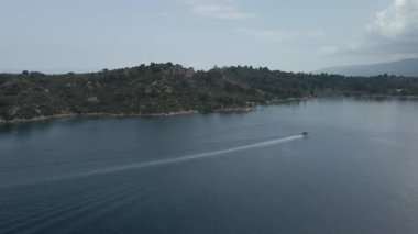 Yunanistan 'ın Chalkidiki yarımadasındaki Akdeniz' de yeşil plantasyonla kayalık sahil etrafındaki sakin sularda yüzen küçük bir tekne boyunca uçmak.